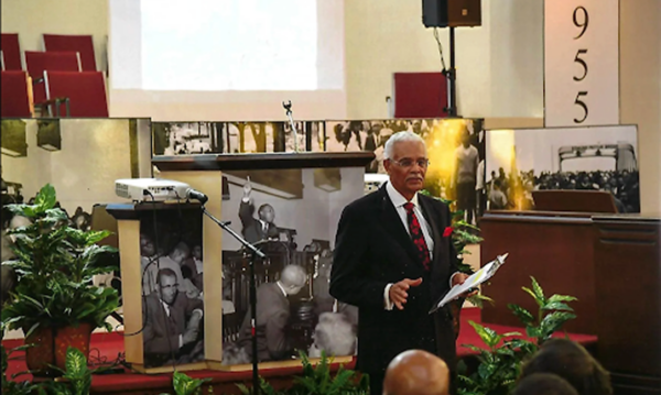 Rev. Willie D. McClung, Ph.D., founder and Chairman of the Board of Holt Street Baptist Church Civil Rights Museum, standing at a podium speaking to a congregation, with historical civil rights photographs displayed on panels behind him.