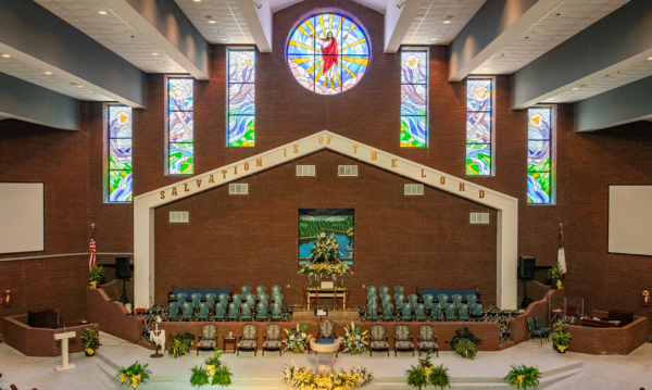 Altar area of Holt Street Memorial Baptist Church, featuring a large round stained-glass window of Christ, flanked by narrower stained-glass panels, with the phrase 'Salvation Is of the Lord' above the choir seating and a floral-decorated altar below.
